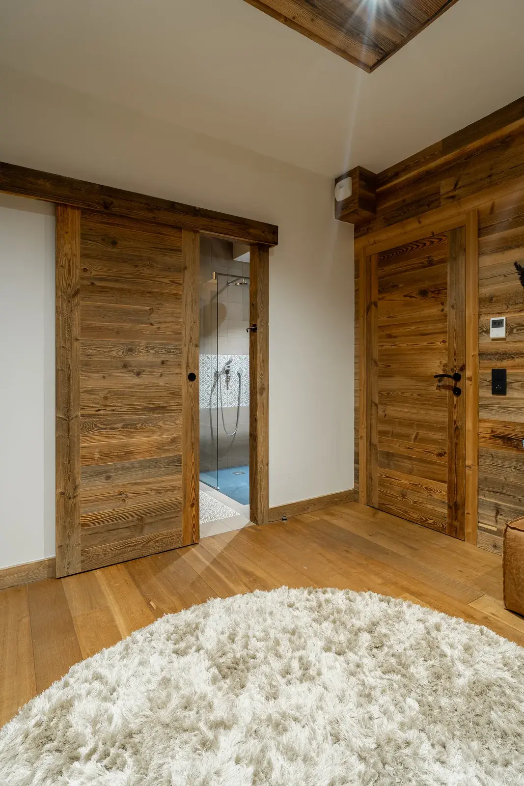 Chambre de chalet : porte coulissante en vieux bois en applique ouverte sur la salle de bains, et bloc-porte battant en vieux bois dans une paroi habillée du même bardage